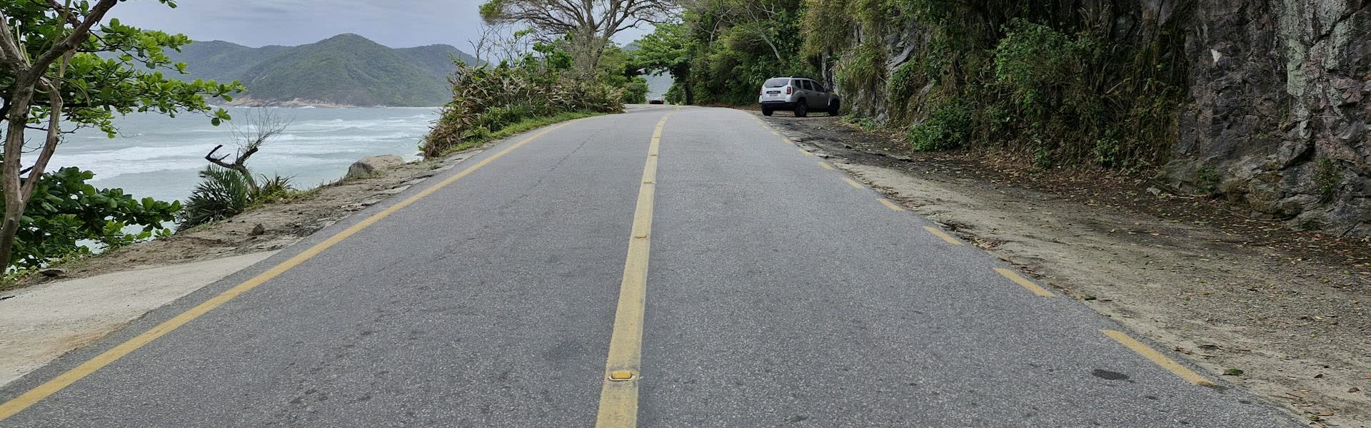 Empty coastal road on an overcast day, ocean to the left and tree-lined cliff to the right.